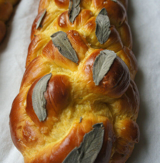 A delicious-looking bright yellow challah decorated with fresh sage leaves lies on a white tablecloth.